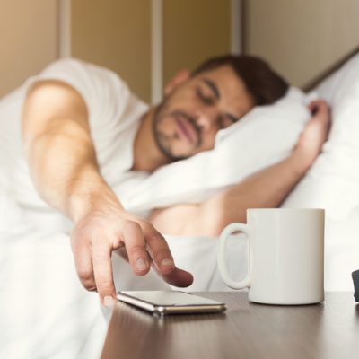 Sleepy guy waking up early after hearing alarm clock signal on smartphone on monday morning, reaching for ringing mobile phone with closed eyes, copy space